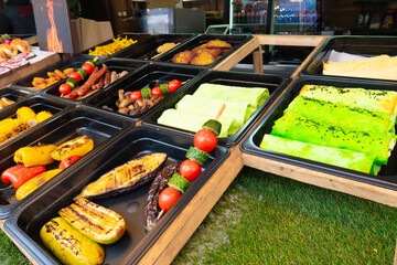 Variety of colorful street food displayed in black trays at a fair, including grilled zucchini, corn, kebabs, and bright green crepes