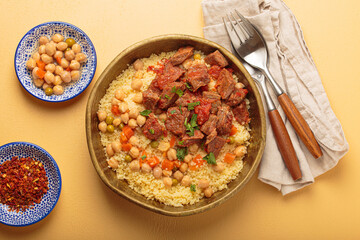 Traditional dish of Maghreb cuisine, a Moroccan couscous with lamb or beef, vegetables, chickpeas, and peas in a rustic bowl with cutlery on yellow background