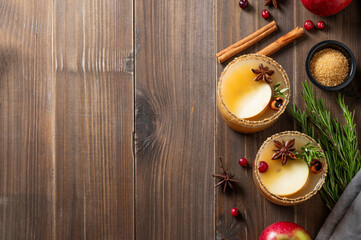 Two glasses of hot apple cider with cinnamon, anise, brown sugar and rosemary on a wooden background