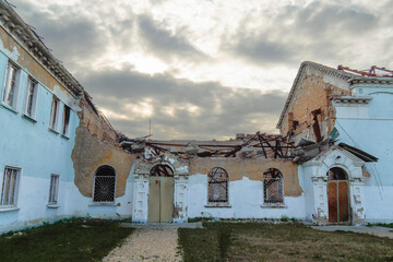 The central section of a historic building is completely destroyed by war, showing a collapsed roof, exposed brick, and empty arched window frames under a dramatic sky