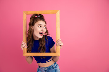 Young woman posing creatively with a wooden frame against a pink background, showcasing trendy...