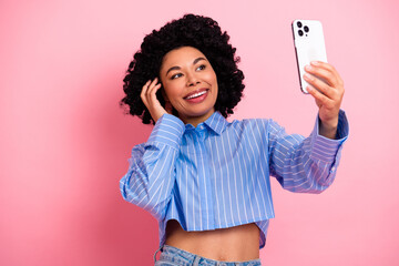 Naklejka na ściany i meble Attractive young woman with stylish curly hair taking a selfie on smartphone against a vibrant pink background