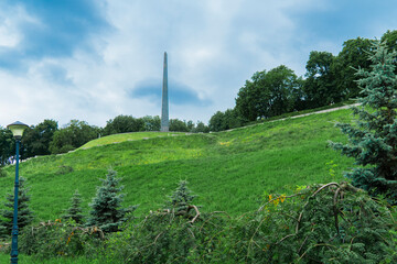 View of the high obelisk monument atop a steep green grassy hill, surrounded by lush park trees under a dramatic blue and white cloudy sky
