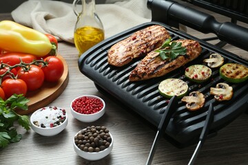Electric grill with tasty chicken fillet, mushrooms, spices and vegetables on wooden table, closeup