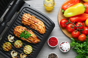 Electric grill with tasty chicken fillet, mushrooms, spices and vegetables on wooden table, flat lay
