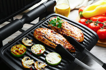 Electric grill with tasty chicken fillet, mushrooms, zucchini and tongs on wooden table, closeup