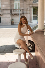 Elegant young woman in chic white dress posing confidently at a stylish outdoor cafe seating area on a sunny day