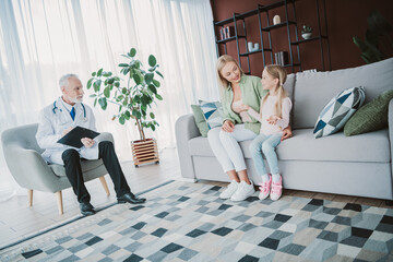 Family scene at home with doctor talking to mother and daughter on sofa in cozy living room