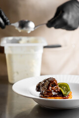 A chef in black gloves plates a gourmet dish of braised beef cheek on a bed of sweet potato puree, garnished with a bell pepper slice on a white plate.