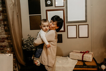 Fototapeta premium Happy grandmother holding and playing with her baby grandson, celebrating a family moment near a decorated Christmas tree in a cozy, rustic holiday home setting. New Year, Christmas