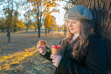 Portrait of a young woman enjoying a coffee and pastry snack in a park, leaning against a tree trunk on a sunny autumn day with yellow leaves