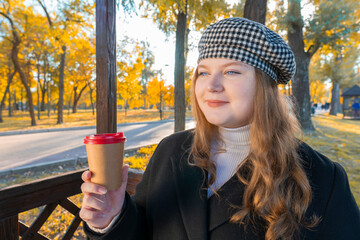 Close-up portrait of a smiling young woman in a checkered beret holding a paper coffee cup in an autumn park with golden foliage