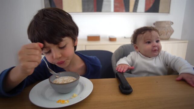 Older brother eating cereal beside baby sibling at breakfast sharing quiet family moment of warmth and innocence in morning light