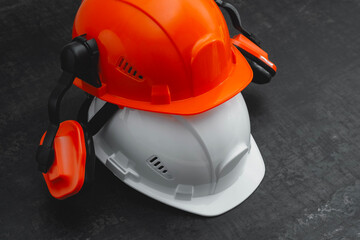 Two construction helmets, orange and white with earmuffs on a dark textured table. Professional...