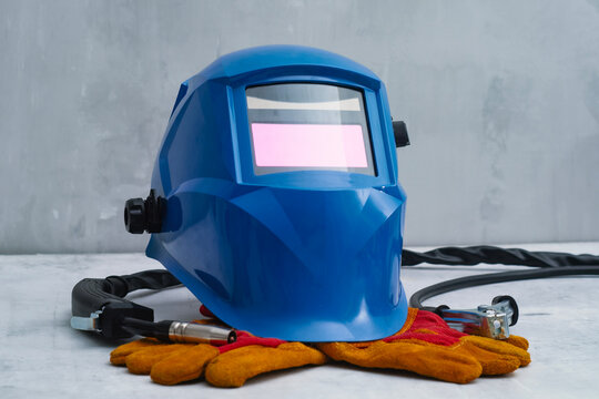 Blue protective welding helmet with orange safety gloves and welding torch placed on a white industrial table. Concept of professional equipment, metalwork and workplace safety.
