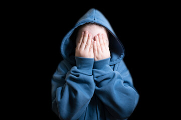 Close-up of a scared or shy child wearing a blue hooded sweatshirt, hiding their face with both hands in front of a black background