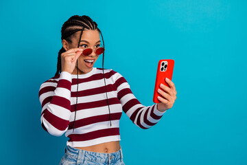 Naklejka na ściany i meble Young stylish woman smiles at her phone wearing a striped top and red sunglass against a bright blue background
