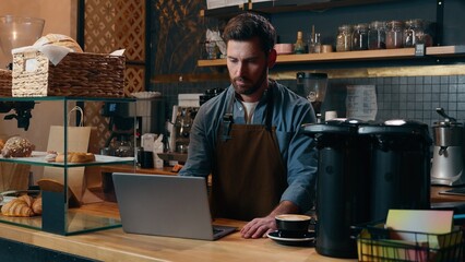 Pensive Caucasian staff employee restaurant worker barista guy using laptop ordering products online shopping in Internet male small business owner thinking in cafeteria waiter man type computer cafe