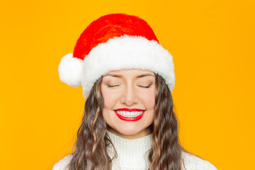 Portrait of young pretty brunette woman wearing Santa hat and a knitted white sweater with red lipstick on her lips closing her eyes and smiling at yellow background.