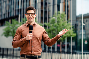 Confident young businessman in urban setting holding microphone during outdoor presentation in...