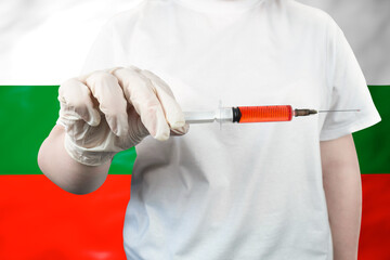 A healthcare professional holding a syringe containing red liquid against the waving national flag of Bulgaria, symbolizing Bulgarian medicine