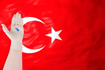 A hand in a protective glove holding a single blue pill against the waving flag of Turkey, symbolizing medical care and health