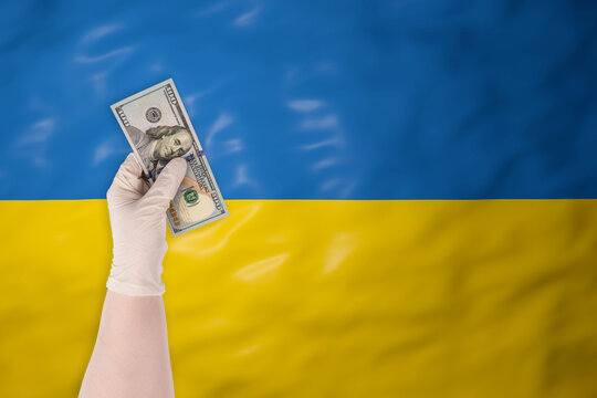 A hand in a protective glove holding a hundred US dollar bill against the background of the waving flag of Ukraine, symbolizing financial aid