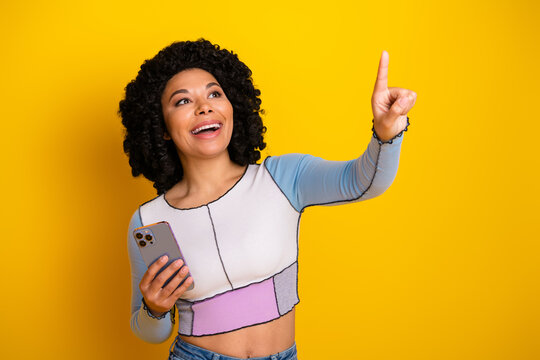Smiling young woman in stylish casual wear pointing upwards with confidence while holding a smartphone against a yellow background - Powered by Adobe