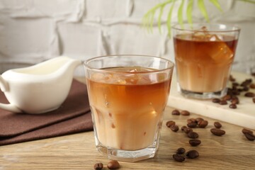Tasty iced coffee with milk in glasses and beans on wooden table against light textured background, closeup