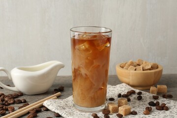 Tasty iced coffee with milk in glass, beans and sugar cubes on wooden table against grey background, closeup