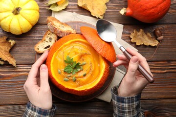 Woman with tasty pumpkin cream soup with seeds and parsley served in pumpkin at wooden table, top...