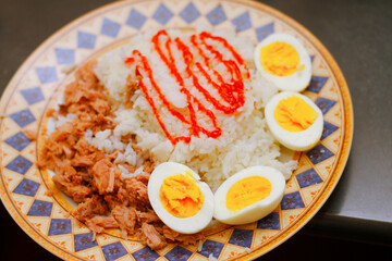 A plate of white rice with shredded meat, red sauce, and hard-boiled eggs is presented on a patterned dish.