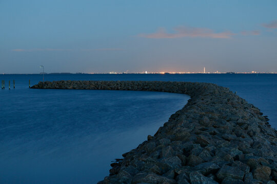 A rocky breakwater curves into the serene blue water under a twilight sky, with distant city lights twinkling on the horizon.