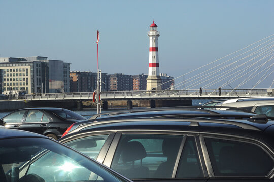 A red and white striped lighthouse stands tall behind a modern bridge where people walk, with parked cars in the foreground and city buildings in the distance.
