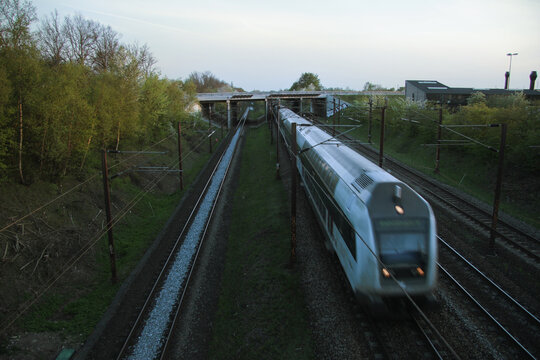 Denmark - A high-speed train blurs as it travels along a track, passing under a bridge in a twilight setting surrounded by green trees.