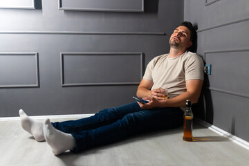 Man sitting against wall looking groggy with a drink in a dimly lit room at night