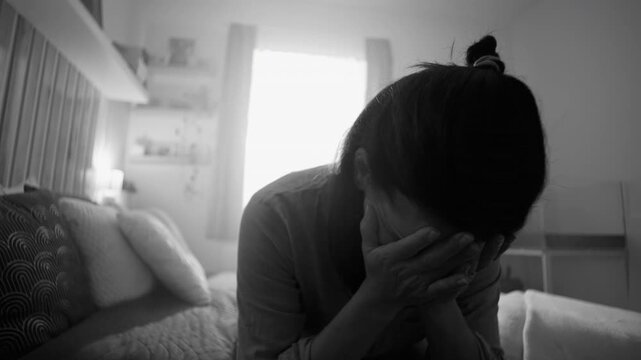 Woman covering face with both hands in silent anguish, expressing sorrow, exhaustion, and emotional breakdown while sitting alone on bed, black and white