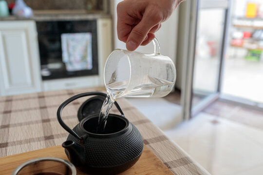 Close-up of a hand pouring hot water from a clear glass jug into a traditional black cast iron teapot (tetsubin) on a wooden board, preparing tea - Powered by Adobe