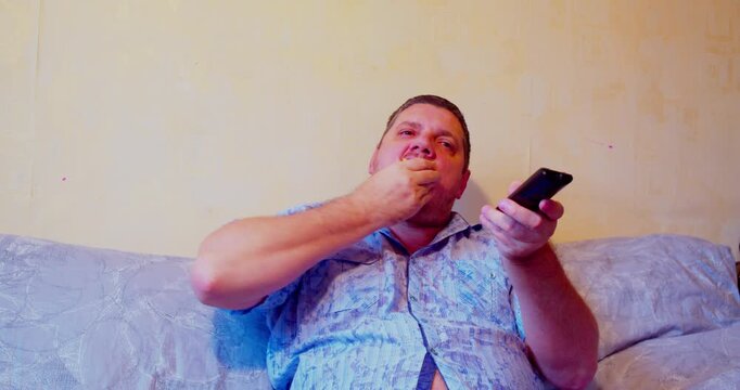 Middle-aged man sitting on couch, eating apple and holding TV remote control, enjoying leisure time at home