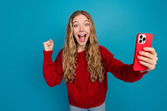 Cheerful young woman holding red smartphone, expressing excitement and joy against vibrant blue background in casual fashion