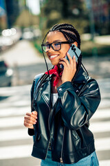 Lovely woman with braided hair holding a smartphone on a city street sidewalk wearing a leather...