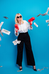 Confident mature businesswoman celebrates success with money toss against blue background wearing...