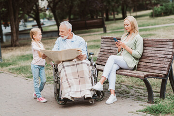 Park moment as family care grandpa in wheelchair reads newspaper while granddaughter chats and...