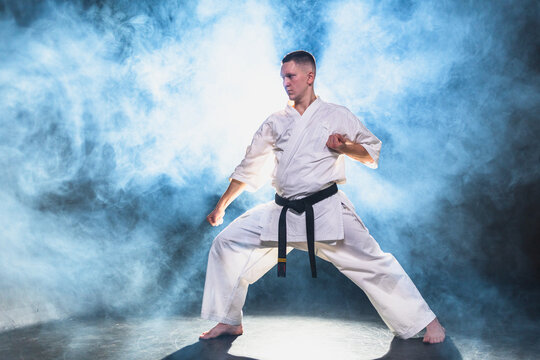 Strong man fighter in white kimono for karate martial arts with black belt preparing for fight training in black background. Man training moves punches kicks practicing shadow fight ready for sparring