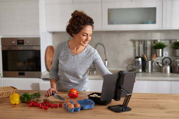 A woman prepares a meal in a sleek kitchen, using a cutting board with vegetables and eggs while following a recipe displayed on a tablet stand.