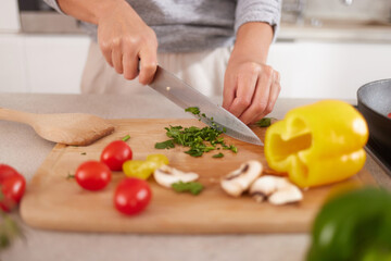 A person is chopping fresh herbs and colorful vegetables like bell peppers, tomatoes, and mushrooms on a wooden cutting board in a bright kitchen setting.
