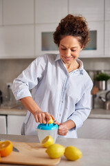 In a sunny kitchen, a woman with curly hair squeezes fresh lemons with a manual juicer. She looks...
