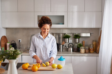 A woman with curly hair cuts lemons on a wooden board in a well-lit kitchen. Fresh oranges are visible on the counter, highlighting a cheerful cooking moment.