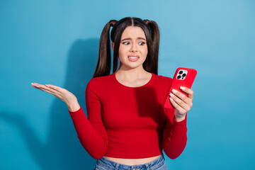 Concerned young woman with ponytails looking at her smartphone against a blue background, wearing a red stylish top