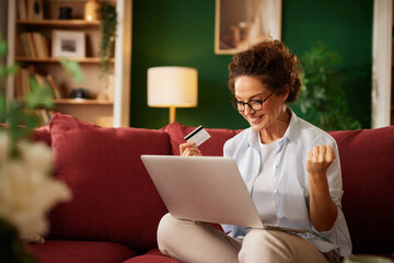 A woman smiles with joy while shopping online. She holds a credit card and has just made a successful purchase. The cozy living room creates a welcoming atmosphere.